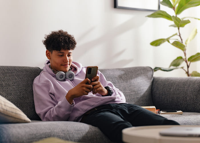 A teenage boy in a purple hoodie and headphones sits on a sofa, using his smartphone in a bright, cozy living room. The scene evokes a sense of modern adolescence and connectivity.