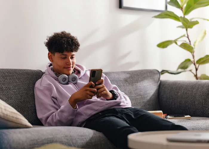 A teenage boy in a purple hoodie and headphones sits on a sofa, using his smartphone in a bright, cozy living room. The scene evokes a sense of modern adolescence and connectivity.