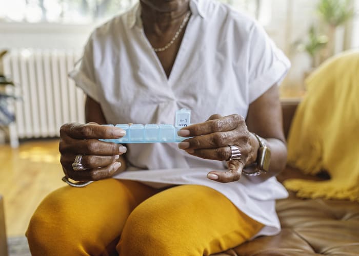 Senior black woman sits on the couch at home and takes medications from a daily pill organizer. Cropped shot does not show the woman's face.