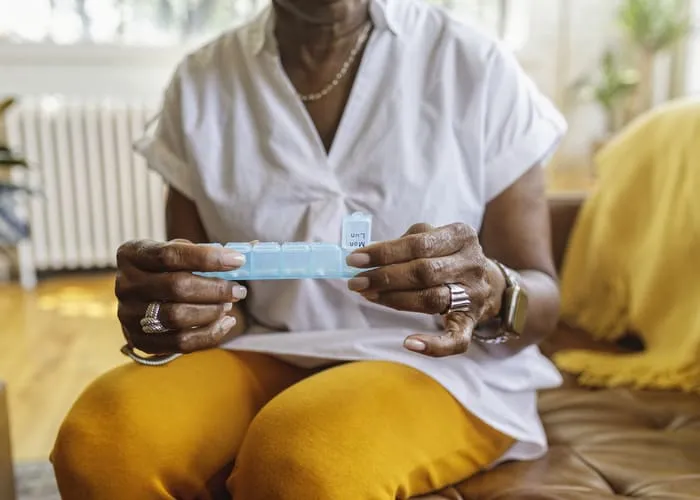 Senior black woman sits on the couch at home and takes medications from a daily pill organizer. Cropped shot does not show the woman's face.